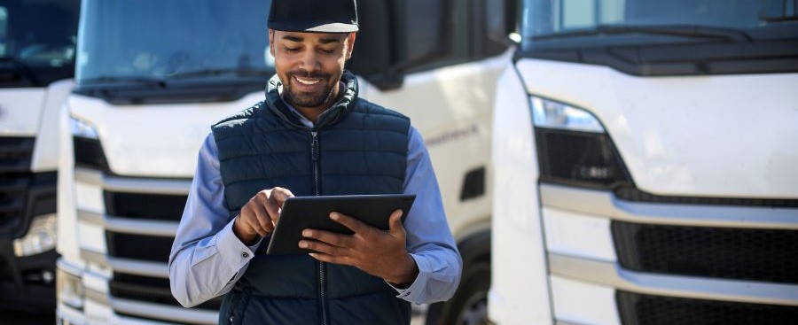 Een afbeelding van een lachende man die een tablet vasthoudt. Achter hem staan vrachtwagens Een afbeelding van een lachende man die een tablet vasthoudt. Achter hem staan vrachtwagens